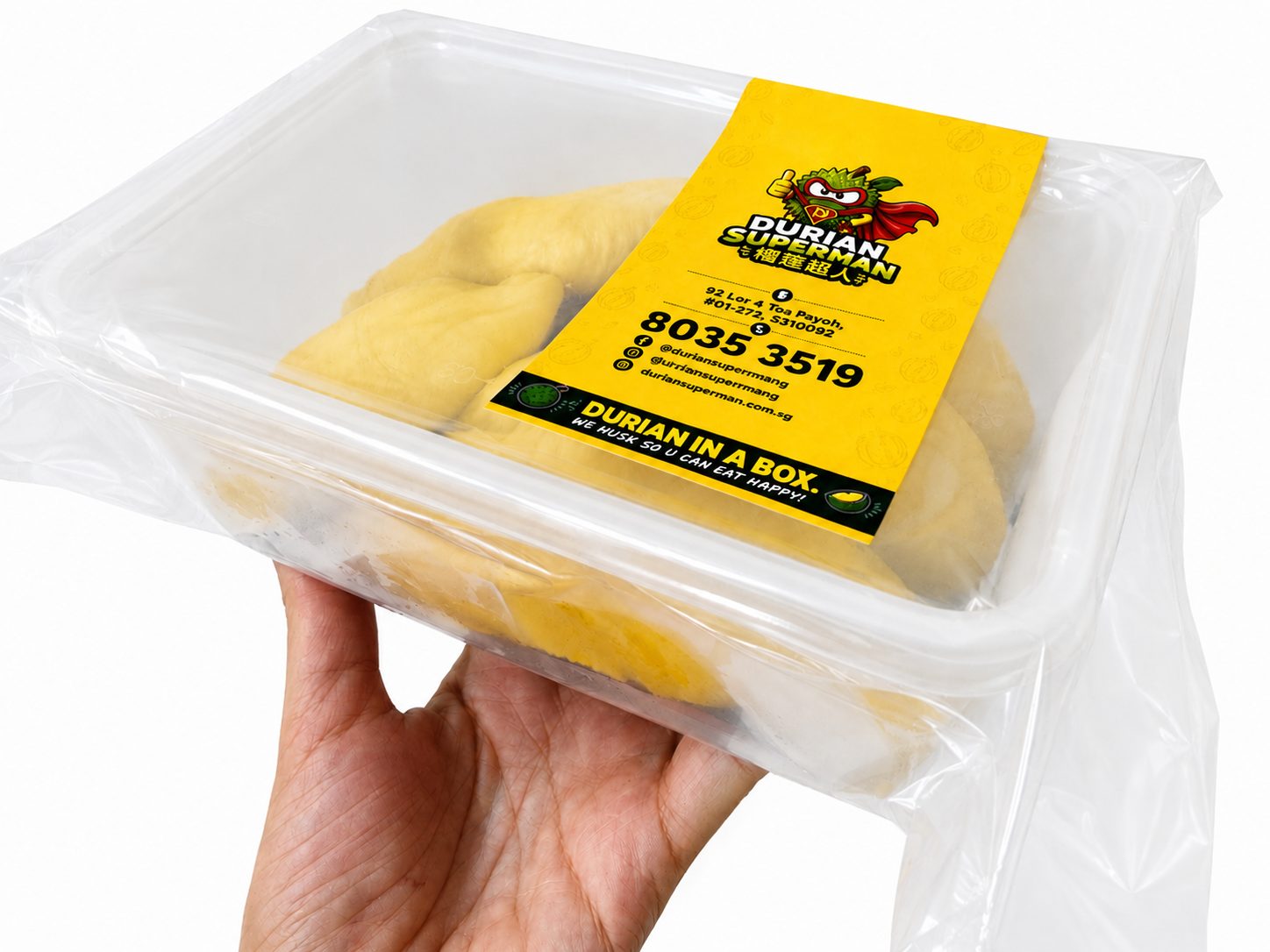 A hand holding a 400g Durian Superman box — dehusked durian flesh vacuum sealed under a yellow label that reads ‘Durian in a box. We husk so u can eat happy!’ with the stall address and phone number